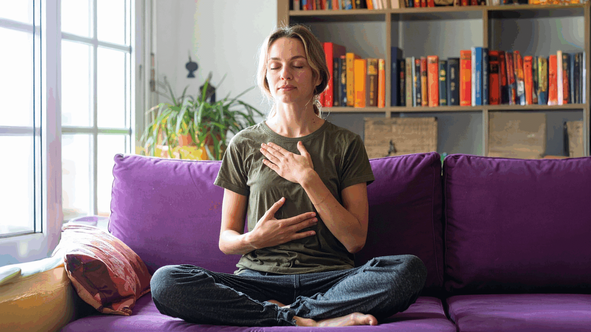 Woman sitting peacefully with hand on heart in a quiet, book-filled room, symbolizing the inner strength and emotional warmth of self-compassion during grief and loneliness.