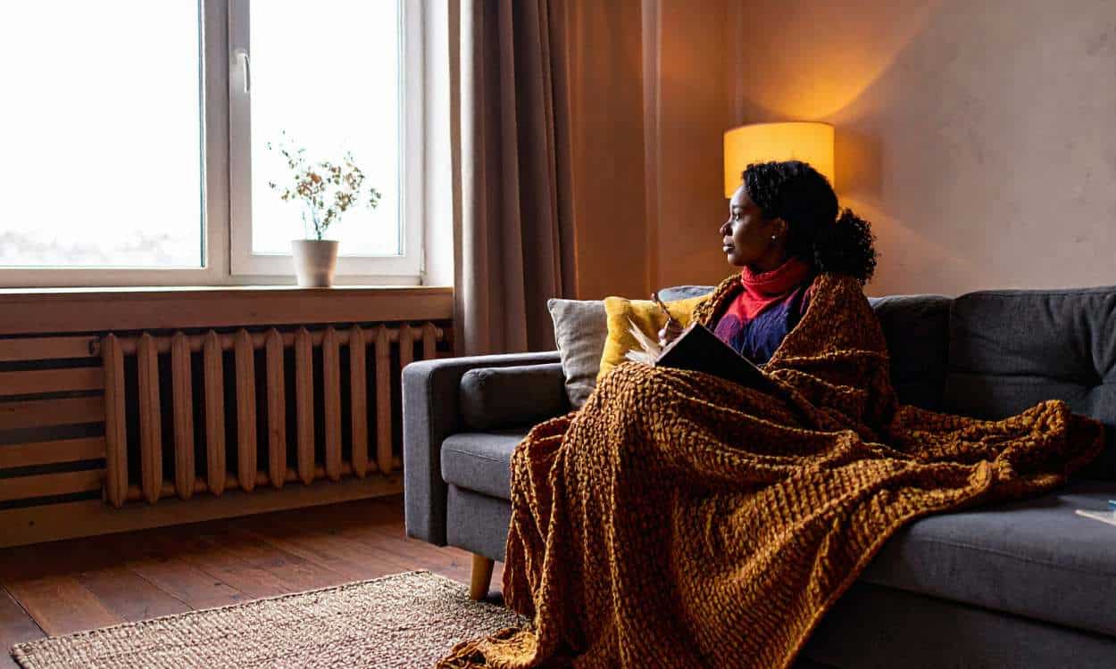 Woman in her 50s sitting alone on a couch with a journal, gazing out the window in quiet reflection, symbolizing grief-related loneliness and emotional isolation.